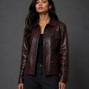 Puede incluir: Una chaqueta de cuero marrón oscuro con cuello clásico, cierre frontal de botones, mangas largas con puños abotonados y dos bolsillos en el pecho con solapas abotonadas. Se lleva sobre una camiseta negra y pantalones cargo oscuros.