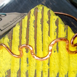 May include: A copper wire bracelet with a wavy design. The bracelet is on a yellow surface with a brown and black background.