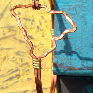 May include: Copper wire bracelet featuring the outline of the Hawaiian Islands. The bracelet has a hammered texture on the island shape and is accented with gold-colored wire wraps. The background is a mix of yellow and blue.