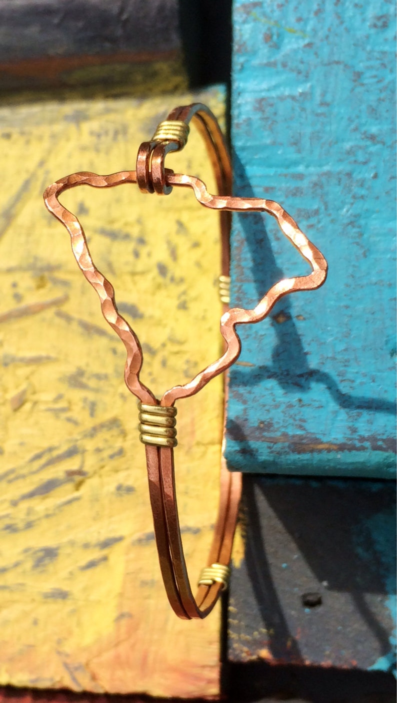 May include: Copper wire bracelet featuring the outline of the Hawaiian Islands. The bracelet has a hammered texture on the island shape and is accented with gold-colored wire wraps. The background is a mix of yellow and blue.