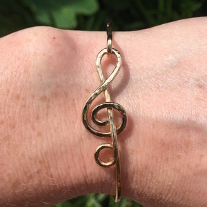 Puede incluir: Un brazalete dorado con un acabado martillado en forma de clave de sol.