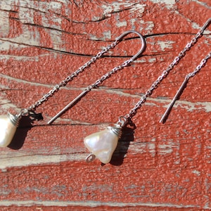 Puede incluir: Un par de pendientes de plata con colgantes en forma de lágrima de color blanco perlado. Los pendientes están colgados en una superficie de madera rústica.