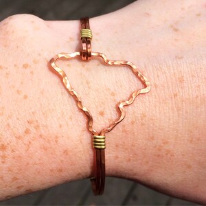 May include: Copper wire bracelet featuring the outline of the state of South Carolina. The bracelet has a hammered texture on the state outline and is connected with gold-colored wire. The bracelet is worn on a wrist.