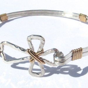 May include: A silver bangle bracelet with a gold cross charm. The bracelet is made of two strands of silver wire, with gold wire wrapped around the strands at the ends. The cross charm is made of silver and has a hammered texture.