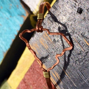 May include: Copper bracelet featuring the outline of the state of South Carolina. The bracelet has a hammered texture and is accented with gold-colored wire. The design is set against a colorful, rustic wooden background.