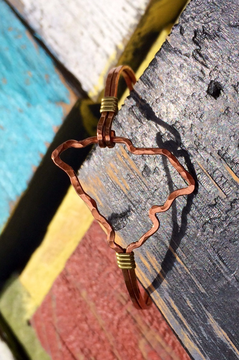 May include: Copper bracelet featuring the outline of the state of South Carolina. The bracelet has a hammered texture and is accented with gold-colored wire. The design is set against a colorful, rustic wooden background.