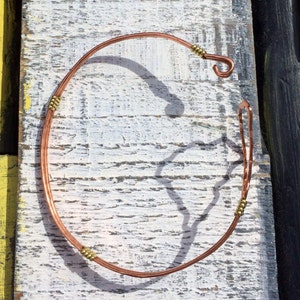 May include: A handcrafted copper necklace with a hook closure. The necklace features small gold-colored beads at intervals along the copper wire. The jewelry is displayed on a weathered, white-painted wooden surface.