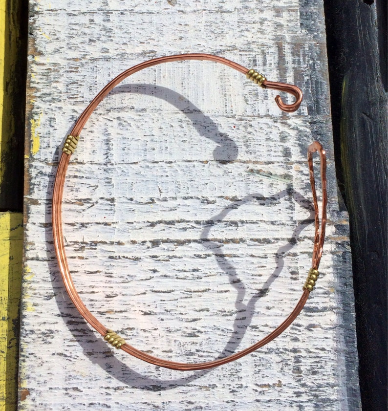 May include: A handcrafted copper necklace with a hook closure. The necklace features small gold-colored beads at intervals along the copper wire. The jewelry is displayed on a weathered, white-painted wooden surface.