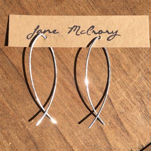 May include: A pair of silver hoop earrings with a simple, modern design. The earrings are made of thin wire and are shaped like a long, thin oval. The earrings are hanging from a brown card with the name "Jane McRory" written on it.