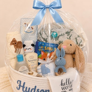 May include: A white rope basket personalised with the name Hudson in blue embroidery, filled with baby items including a tan plush rabbit, a blue crochet rattle, Aveeno baby lotion, a blanket with a bear, a dinosaur sleeper, and a book.