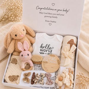 May include: A white gift box containing a tan plush bunny, a pink crocheted bunny rattle, a white onesie with 'hello world' text, a bear-patterned muslin cloth, a wooden birth announcement disc, hair clips, and a floral accessory set.