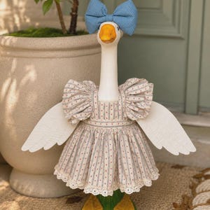 May include: A white goose statue wearing a blue polka dot bow on its head and a beige dress with vertical blue stripes and small red dots. The dress features ruffled sleeves and a white lace trim along the bottom hem, covering the goose's torso.