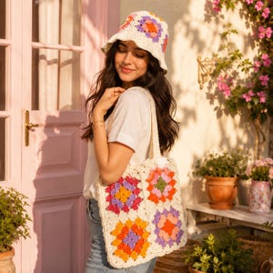 Op de afbeelding: Een gehaakte bucket hat en een bijpassende tas, beide met een witte basis en kleurrijke granny square motieven in tinten oranje, roze, paars en groen. De items zijn gemaakt van garen met een getextureerd, handgemaakt uiterlijk.