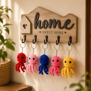 Könnte beinhalten: Ein hölzerner Schlüsselhalter für die Wand mit dem Schriftzug home und SWEET HOME sowie fünf schwarzen Metallhaken. An den Haken hängen fünf gehäkelte Oktopus-Schlüsselanhänger in Rot, Hellrosa, Blau, Dunkelrosa und Gelb mit schwarzen Augen.