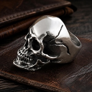 May include: A silver-toned metal ring shaped like a human skull. The ring features a polished finish with recessed dark areas to highlight the anatomical details of the eye sockets, nasal cavity, and teeth. The skull design forms the main band.