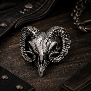 May include: A silver-toned metal ring featuring a detailed ram skull design with prominent, textured curved horns. The surface of the ring has a weathered, oxidized finish that highlights the intricate grooves and contours of the skull structure.