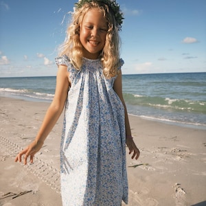 May include: A sleeveless dress featuring a blue and white floral pattern with a smocked neckline and ruffled shoulder details. The garment has a loose, flowing silhouette that falls to the mid-calf, crafted from a lightweight fabric.