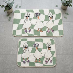 Peut inclure: Deux tapis de bain rectangulaires avec un motif à carreaux blanc et vert sauge. Chaque tapis présente trois canards blancs : les deux extérieurs portent des serviettes blanches sur la tête et des lunettes, le central porte des bigoudis violets.