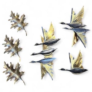May include: A collection of metal wall art pieces featuring three oak leaves with a metallic gold and dark finish, a vertical grouping of three flying geese, a pair of geese in flight, and a single goose in flight, all with gold and dark tones.
