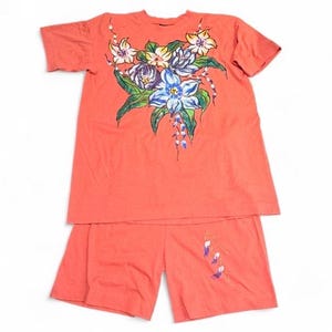 May include: A coral-colored short-sleeve t-shirt and matching shorts set. The t-shirt features a hand-painted floral design in shades of blue, yellow, and green on the chest. The shorts include a small, delicate floral accent on the lower right leg.
