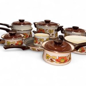 May include: A collection of white enamel cookware featuring brown lids and handles with a floral pattern in orange, yellow, and green. The set includes various pots, pans, and a small cup, all decorated with the same botanical motif.