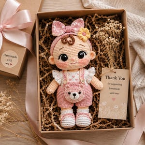 Peut inclure: Une poupée au crochet avec de grands yeux noirs, portant une salopette rose à motif d'ours, un chemisier blanc à volants et un bandeau à nœud rose avec une fleur jaune. La poupée est dans une boîte cadeau avec du papier froissé et une étiquette.