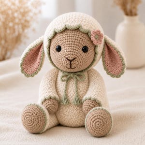 May include: A crocheted stuffed animal shaped like a lamb with a cream-colored body, tan paws, and large black eyes. The lamb wears a cream bonnet with sage green scalloped trim, a small pink flower accent, and a sage green tie with tassels.