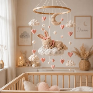 Può includere: Una giostrina sospesa per la cameretta con una cornice circolare in legno e elementi all'uncinetto, tra cui un coniglietto che dorme su una nuvola, una luna crescente, nuvole bianche e piccoli cuori rosa appesi a fili sottili.