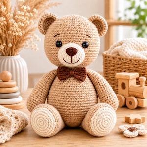 Könnte beinhalten: Ein hellbrauner, gehäkelter Teddybär mit einer dunkelbraun gestickten Nase und einer passenden dunkelbraunen Fliege. Der Bär hat runde Ohren, schwarze Kunststoffaugen und hellbeige, kreisförmige Applikationen an den Fußsohlen aus Garn.
