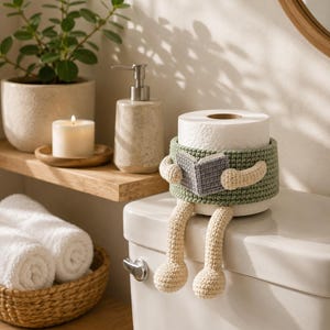 May include: A crochet toilet paper roll cover featuring a sage green band with attached cream-colored dangling legs and arms. The figure holds a small grey open book in its hands, wrapped around a standard white roll of toilet paper.
