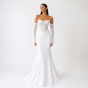Könnte beinhalten: Ein bodenlanges weißes Spitzenbrautkleid mit einem trägerlosen Korsett-Oberteil mit sichtbaren Stäbchen und passenden abnehmbaren langen Spitzenärmeln. Das Kleid hat eine figurbetonte Silhouette, die in einer Schleppe ausläuft.