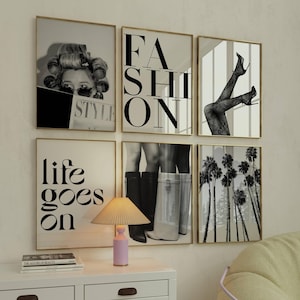 May include: A gallery wall set of six framed black and white prints featuring fashion-themed imagery, including text that reads FASHION and life goes on, alongside a small table lamp with a pleated beige shade and a light pink cylindrical base.
