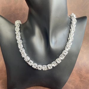 May include: A silver-toned necklace featuring a thick, interlocking Byzantine chain link design. The chain is composed of multiple small, circular metal links woven together to create a continuous, textured, and cylindrical rope-like pattern.