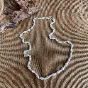 May include: A silver-toned chain necklace featuring a series of repeating, textured, barrel-shaped links. The necklace is constructed with a metallic finish and includes a small, visible hook-style clasp mechanism at the top of the chain loop.