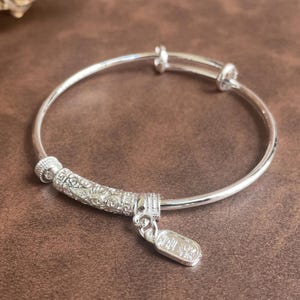May include: A silver-toned bangle bracelet featuring a smooth, thin circular band with an adjustable overlapping mechanism. It includes a textured, cylindrical bead with intricate engraved patterns and a small, rectangular dangling charm.