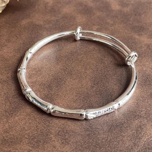 May include: A silver-toned expandable bangle bracelet featuring a segmented design that mimics the appearance of bamboo stalks. The bracelet has a polished metallic finish and includes a small section adorned with tiny, clear, sparkling stones.