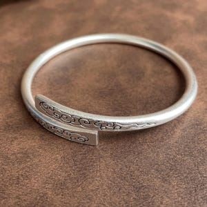 May include: A silver-toned open cuff bracelet with a matte finish. The bracelet features a rounded band that tapers into two flat, overlapping ends. Each end is engraved with a detailed, repeating cloud pattern that wraps around the metal surface.