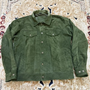 Puede incluir: Una chaqueta de ante verde oliva con cuello clásico, cierre frontal de botones metálicos, dos bolsillos en el pecho con solapas abotonadas y mangas largas con puños abotonados. La chaqueta presenta un corte estructurado con costuras verticales.