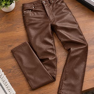 May include: A pair of dark brown trousers with a smooth, leather-like finish. The trousers feature a classic five-pocket design, a button and zip fly closure, and silver-toned metal rivets at the pocket corners, showcasing a straight-leg cut.