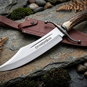 May include: A fixed-blade knife with a polished steel blade featuring the text CUTTINGEDGEBLADE, a textured brown antler handle, and a dark metal crossguard. The knife is accompanied by a brown leather sheath with a strap and metal rivet.