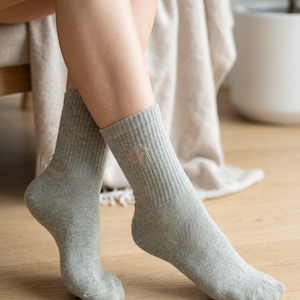 May include: A pair of light gray ribbed crew socks featuring a small, embroidered gold-colored logo on the outer side of the ankle. The socks have a textured knit pattern and reach mid-calf, providing a simple and functional design for everyday wear.
