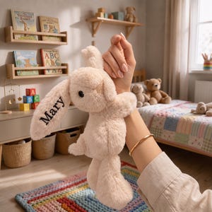 May include: A plush cream-colored stuffed rabbit toy with long ears, featuring the name Mary embroidered in black thread on one ear. The rabbit has a soft, fuzzy texture, dark round eyes, and a small stitched nose, designed as a cuddly companion.