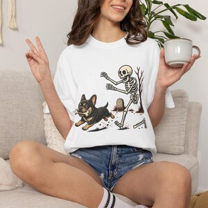 May include: A white short-sleeve t-shirt featuring a graphic print of a black and tan French Bulldog running with a bone in its mouth, being chased by a cartoon skeleton in a graveyard setting with a tombstone and a bare tree in the background.