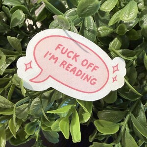 May include: A white die-cut sticker shaped like a speech bubble with a pink outline. The sticker features the text FUCK OFF I'M READING in pink capital letters, accompanied by two small pink four-pointed star icons on each side of the text.