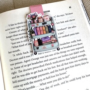 May include: A magnetic bookmark featuring a light blue three-tiered rolling cart illustration. The cart holds books, a candle, a vase of flowers, a mug, and a black cat. The bookmark has a pink top tab and is placed on an open book page.