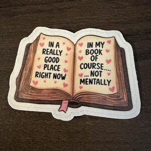 May include: A die-cut sticker featuring an open book with brown covers and a small pink bookmark. The pages are cream-colored with small red hearts and black text that reads: IN A REALLY GOOD PLACE RIGHT NOW IN MY BOOK OF COURSE.... NOT MENTALLY.
