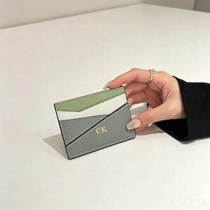 Puede incluir: Un tarjetero rectangular con un diseño de bloques de color en verde salvia, blanco y gris pizarra. El panel frontal incluye un bolsillo diagonal con las iniciales CK grabadas en oro en el centro de la sección gris. Bordes cosidos.