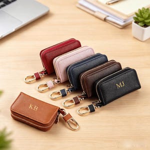 May include: A collection of six rectangular zippered key cases in red, light pink, navy blue, dark brown, and black. Each case features a gold-toned metal lobster clasp and a zipper closure. Two cases are embossed with the gold initials KB and MJ.
