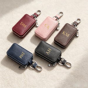 May include: Five rectangular textured key cases with silver-tone zippers and metal swivel clips. The cases come in burgundy, light pink, brown, navy blue, and black, each featuring gold-tone embossed initials: DM, CK, KM, DM, and HY respectively.