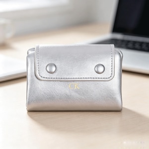 May include: A metallic silver bifold wallet featuring a flap closure secured by two matching silver snap buttons. The front of the wallet displays the gold-colored letters CK embossed in the center, providing a subtle contrast to the smooth finish.